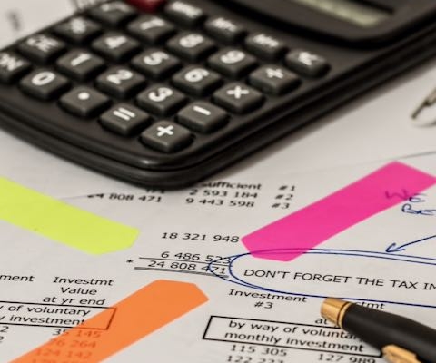 Das Foto zeigt eine Kostenaufstellung, ein gelbes, ein orangenes und ein pinkes, schmales Post-it, die auf eine bestimmte Zahl hinweisen und die Aufmerksamkeit des Betrachters auf diese Zahl lenken sollen. Ein schwarz-goldener Kugelschreiber deutet ebenfalls auf diese Zahl. Im Hintergrund, liegt, leicht verschwommen, ein schwarzer Taschenrechner.