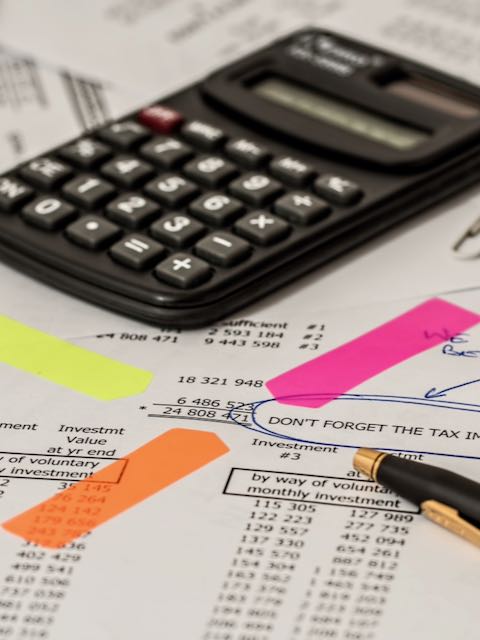 Das Foto zeigt eine Kostenaufstellung, ein gelbes, ein orangenes und ein pinkes, schmales Post-it, die auf eine bestimmte Zahl hinweisen und die Aufmerksamkeit des Betrachters auf diese Zahl lenken sollen. Ein schwarz-goldener Kugelschreiber deutet ebenfalls auf diese Zahl. Im Hintergrund, liegt, leicht verschwommen, ein schwarzer Taschenrechner.