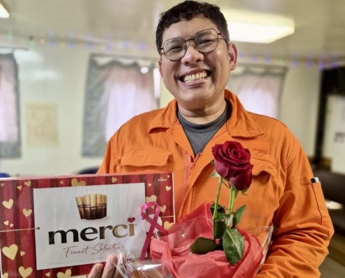 Grüße zum Valentinstag an Bord eines Schiffes im Hafen von Le Havre. Ein philippinischer Seemann hält eine Packung Schokoalde und eine rote Rose lächelnd in de Kamera.