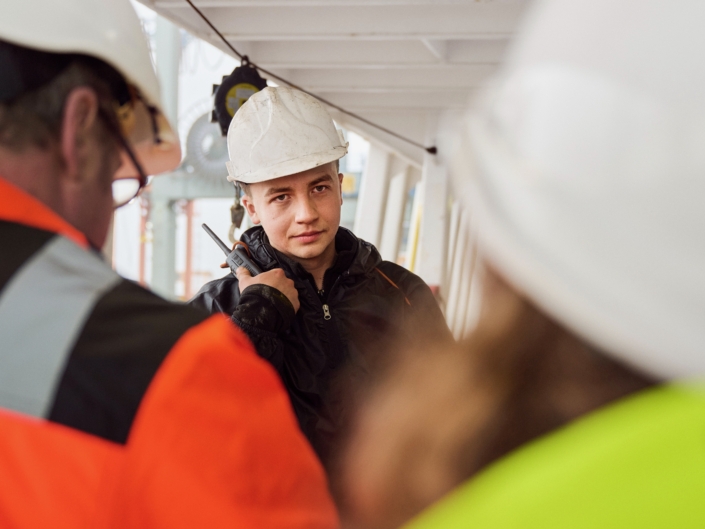 Bordbesuche, wie hier symbolisch dargestellt, sind enorm wichtig für Seeleute, weil diese häufig nicht an Land können oder dürfen. Das Foto zeigt einen jungen Seemann an Bord eines Schiffes. Er trägt einen weißen Schutzhelm, Arbeitskluft und hat die rechte Hand an einem schwarzen Funkgerät. Mit dem Rücken zum Fotografen stehen zwei Bordbesucher der Deutschen Seemannsmission, links ein Mann mit Brille und rechts eine kleinere Person, sehr wahrscheinlich eine junge Frau mit langen Haaren.