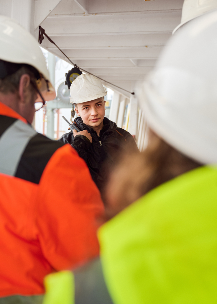 Bordbesuche, wie hier symbolisch dargestellt, sind enorm wichtig für Seeleute, weil diese häufig nicht an Land können oder dürfen. Das Foto zeigt einen jungen Seemann an Bord eines Schiffes. Er trägt einen weißen Schutzhelm, Arbeitskluft und hat die rechte Hand an einem schwarzen Funkgerät. Mit dem Rücken zum Fotografen stehen zwei Bordbesucher der Deutschen Seemannsmission, links ein Mann mit Brille und rechts eine kleinere Person, sehr wahrscheinlich eine junge Frau mit langen Haaren.