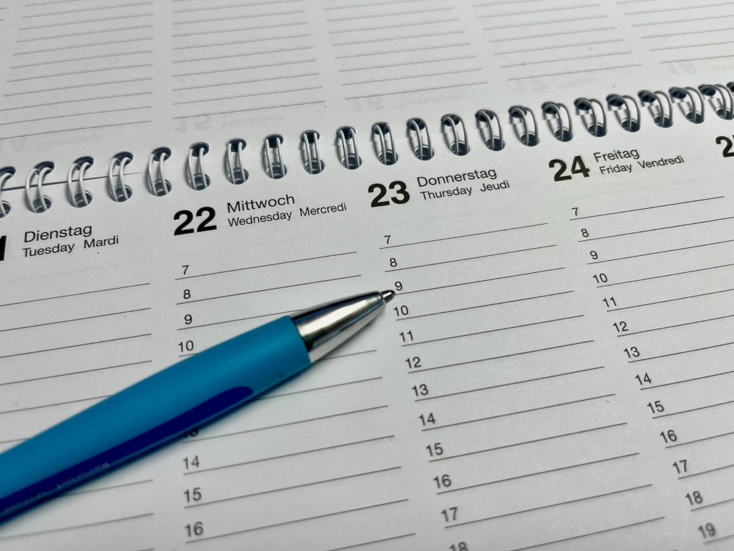 Das Symbolfoto zeigt einen Ausschnitt eines Wochenkalenders, der auf vielen Büroschreibtischen liegen könnte. Ein Kugelschreiber in hellerem und dunklerem Blau gehalten zeigt mit gezückter Miene auf den 23. eines Monats, auf 9 Uhr. Der Wochenkalender hat eine silberne Spiralbindung.