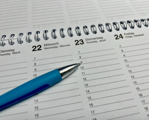 Das Symbolfoto zeigt einen Ausschnitt eines Wochenkalenders, der auf vielen Büroschreibtischen liegen könnte. Ein Kugelschreiber in hellerem und dunklerem Blau gehalten zeigt mit gezückter Miene auf den 23. eines Monats, auf 9 Uhr. Der Wochenkalender hat eine silberne Spiralbindung.