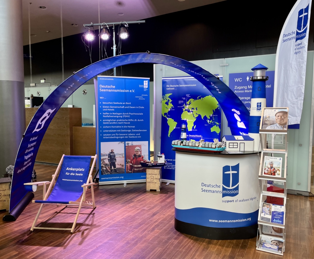 Das Foto zeigt den Messestand der Deutschen Seemannsmission samt dunkelblauem Bannerbogen. Unter dem Bogen sieht man einen leeren Liegestuhl. Rechts im Bild steht der Messestand, daneben ein Aufsteller mit Magazinen und Flyern der Deutschen Seemannsmission.