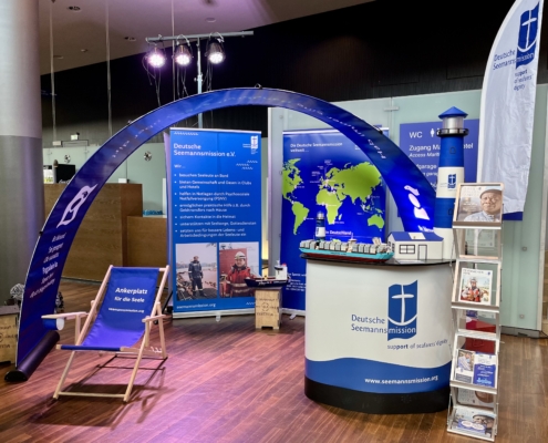 Das Foto zeigt den Messestand der Deutschen Seemannsmission samt dunkelblauem Bannerbogen. Unter dem Bogen sieht man einen leeren Liegestuhl. Rechts im Bild steht der Messestand, daneben ein Aufsteller mit Magazinen und Flyern der Deutschen Seemannsmission.