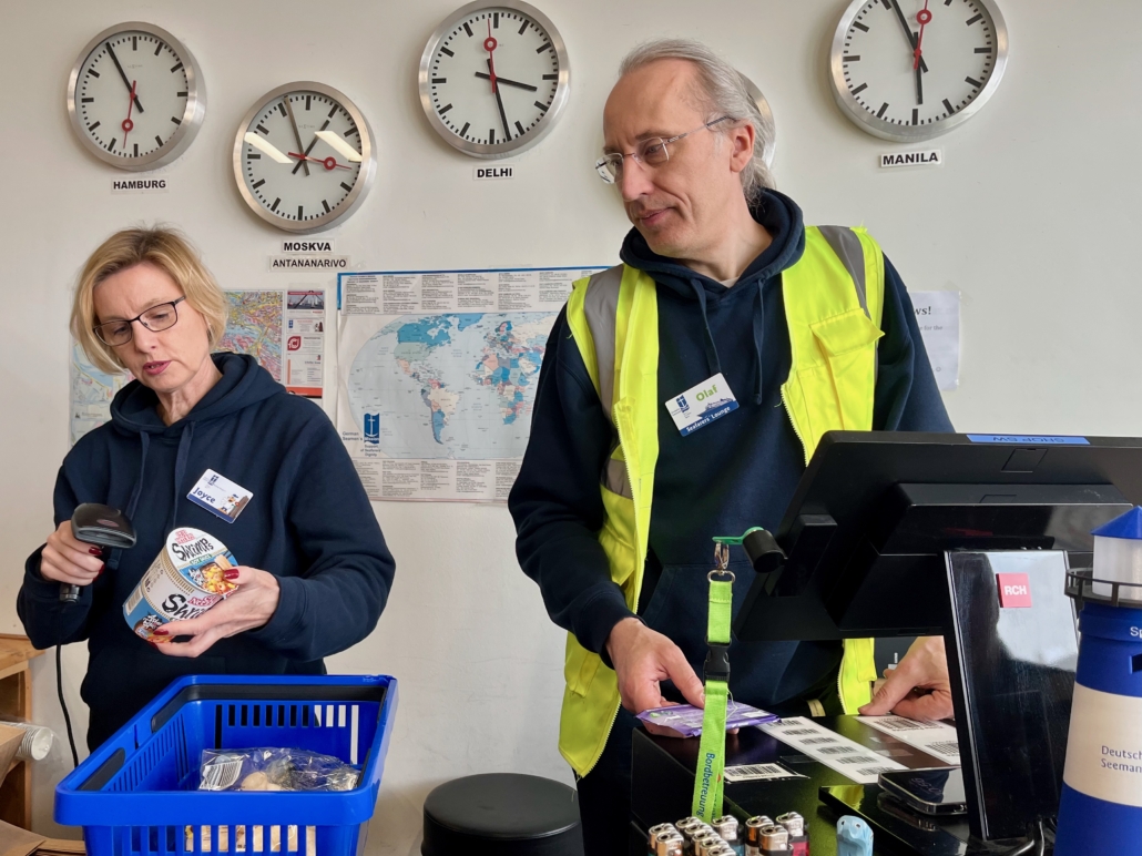 Das Foto zeigt eine Ehrenamtliche und den Leiter der Seafarers' Lounge, Olaf Schröder, an der Kasse der Lounge in Hamburg-Steinwerder. Die Lounge liegt im Kreuufahrtterminal und ist für die Crews der Kreuzfahrtschiffe gedacht. Hinter den beiden an der Wand hängen Uhren mit unterschiedlichen Uhrzeiten und eine Weltkarte. Beide tragen Namensschilder auf ihren dunkelblauen Hoogies. Olaf Schröder trägt auch eine neongelbe Warnweste.