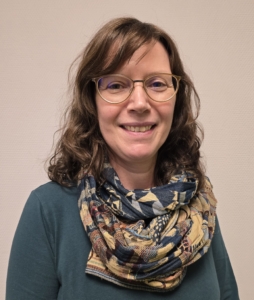 Das Foto zeigt Susanne Frosch, die den Standort der Deutschen Seemannsmission in Hongkong leitet. Sie trägt einen flaschengrünen, langärmeligen Pullover und ein in Beige- und Grüntönen gehaltetnes Halstuch. Ihre schulterlangen braunen Haare, trägt Susanne Frosch offen. Auf ihrem Nasenrücken sitzt eine Brille. Susanne Frosch lächelt freundlich in die Kamera. Sie steht vor einem neutralen, beigfarbenen Hintergrund.