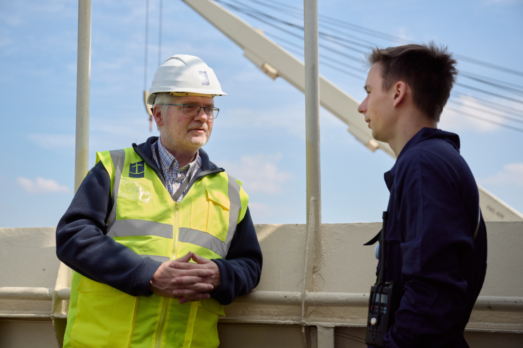 Das Fot zeigt Seemannspastor und Stationsleiter Marc Schippers (li.) mit weißem Schutzhelm und neopngelber Warnweste an Bord eines Schiffes im Hafen von Antwerpen. Er ist im Gespräch mit einem jungen Seemann. Der Mann trägt einen dunkelblauen Arbeitsoverall. Schippers hört dem jungen Seemann aufmerksam zu. 