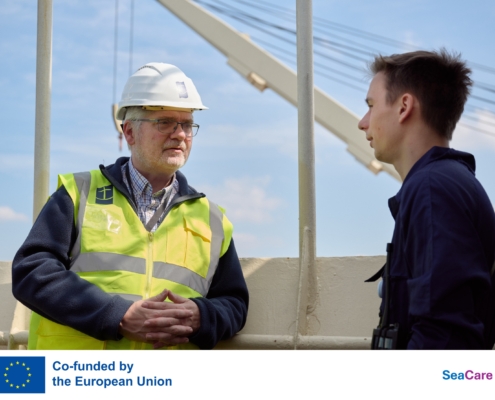 Das Fot zeigt Seemannspastor und Stationsleiter Marc Schippers (li.) mit weißem Schutzhelm und neopngelber Warnweste an Bord eines Schiffes im Hafen von Antwerpen. Er ist im Gespräch mit einem jungen Seemann. Der Mann trägt einen dunkelblauen Arbeitsoverall. Schippers hört dem jungen Seemann aufmerksam zu. Am unteren Rand stehen in einem weißen Kästchen das Logo der EU, mit einem Hinweis, dass das Projekt von der EU gefördert ist und das Sea Care-Logo in Blau-Magenta.