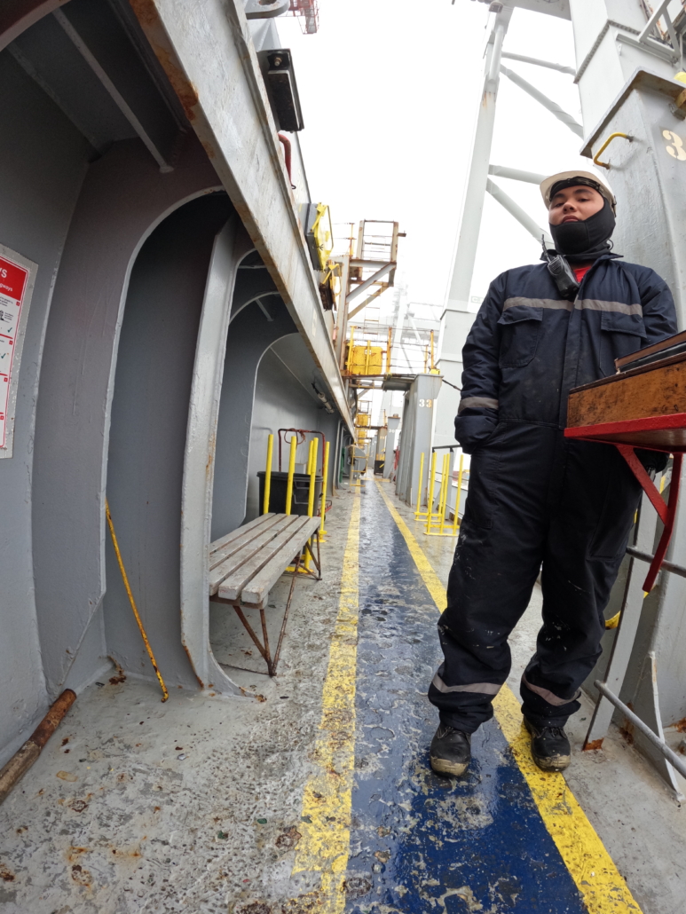 Das Foto ist an Bord eines Containergeschiffs augenommen worden. Man sieht einen philippinischen Seemann, der Watchman, Wachgänger ist. Er begrüßt alle Menschen, die das Schiff betreten möchten und lässt sie sich in eine Liste eintragen. Er steht am rechten Bildrand in seinem dunkelblauen Arbeitsoverall mit weißem Schutzhelm auf dem Kopf und blickt auf die Fotografin hinunter. Der junge Mann ist vom Stahl des Schiffes umgeben. Sein Gesicht ist schwer zu deuten, er wirkt weder fröhlich noch besonders traurig.