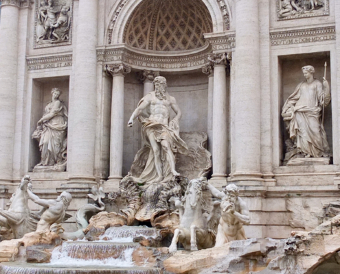 Das Foto zeigt den berühmten Trevibrunnen in Italiens Hauptstadt Rom. Man sieht die kunstvoll gestalteten Figuren, deren Gewänder aus dem Stein gearbeitet sind. Das türkisfarbene Wasser fließt in das Brunnenbecken hinein.