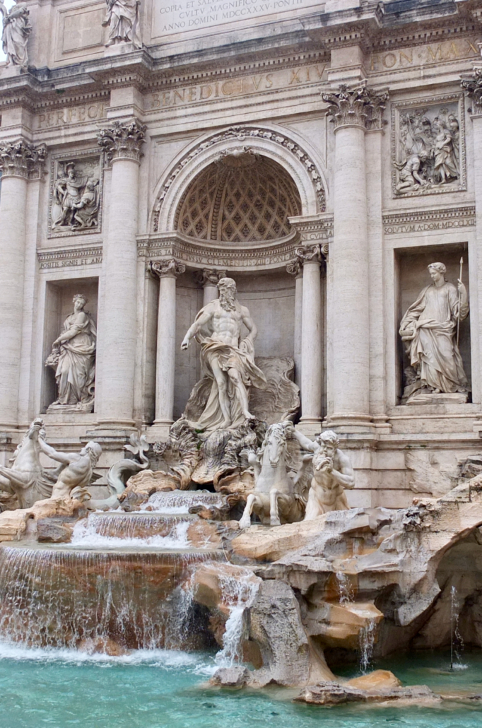 Das Foto zeigt den berühmten Trevibrunnen in Italiens Hauptstadt Rom. Man sieht die kunstvoll gestalteten Figuren, deren Gewänder aus dem Stein gearbeitet sind. Das türkisfarbene Wasser fließt in das Brunnenbecken hinein.